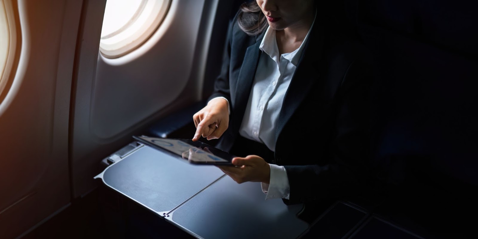 mujer de negocios en el interior de un avión. Está sentada junto a una ventana, utilizando una tablet colocada sobre la mesa desplegable del asiento.